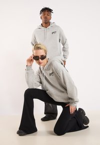 Two models wearing matching grey hoodies with a logo. The first model kneels, wearing black ribbed pants and dark shoes; the second stands behind.