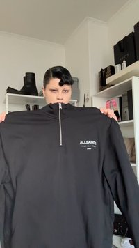 Must kootud pluus kõrge kraega ja poole lukuosaga. Rinnal valge "ALLSAINTS" logo. Sile kangatekstuur, lõdvestunud istuvus.