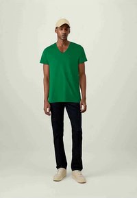 Πράσινο βαμβακερό t-shirt με λαιμόκοψη σε σχήμα V, κοντά μανίκια, με μικρό λογότυπο στο στήθος. Συνδυάζεται με σκούρο τζιν και μπεζ αθλητικά παπούτσια. Ουδέτερο φόντο.