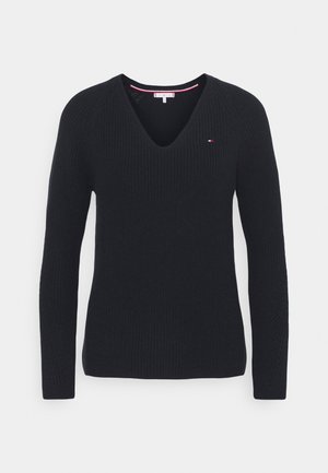 Suéter negro de punto con cuello en V y mangas largas, con un pequeño logo rojo, blanco y azul en el lado izquierdo del pecho, sobre un fondo blanco.