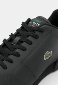 Basket noire Lacoste avec lacets noirs, logo crocodile vert sur le côté et étiquette de marque sur la languette.
