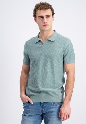 Young man wearing light green short-sleeve textured polo shirt and blue jeans, standing with one hand in pocket, looking to the side.