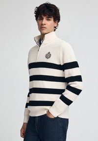 Cremfarbener und marineblauer gestreifter Pullover mit einem halben Reißverschluss am Kragen und einem gestickten Logo auf der Brust, aus strukturiertem Strickmaterial gefertigt.