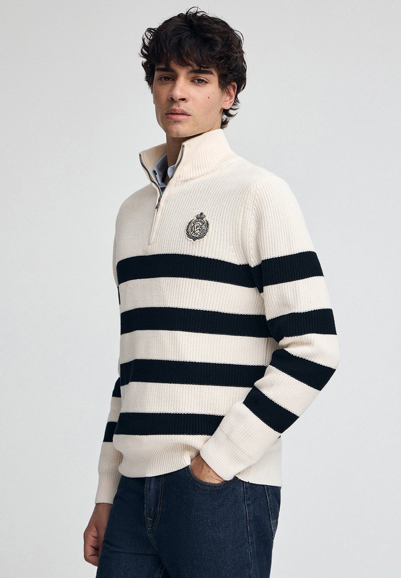 Cremfarbener und marineblauer gestreifter Pullover mit einem halben Reißverschluss am Kragen und einem gestickten Logo auf der Brust, aus strukturiertem Strickmaterial gefertigt.