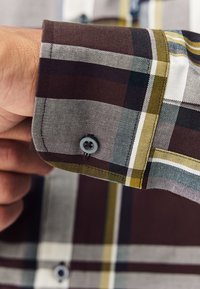 Polsino di camicia a quadri in tessuto marrone, giallo e bianco; presenta una chiusura con bottone e una texture liscia. Vista ravvicinata dei motivi del tessuto.