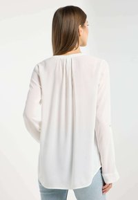 Blusa bianca a maniche lunghe con una vestibilità rilassata, pieghe sottili nella parte posteriore e polsini abbottonati, abbinata a denim azzurro chiaro.