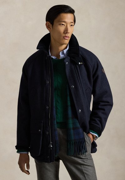 Giacca blu navy in misto lana con collo alto, bottoni a pressione e tasche laterali, indossata sopra un maglione verde lavorato a trecce e una sciarpa a quadri.