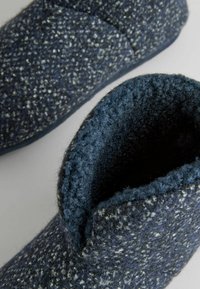 Chausson bleu foncé en laine texturée, doté d'une doublure douce et duveteuse, et d'un design ajusté à la cheville avec un bord légèrement relevé.