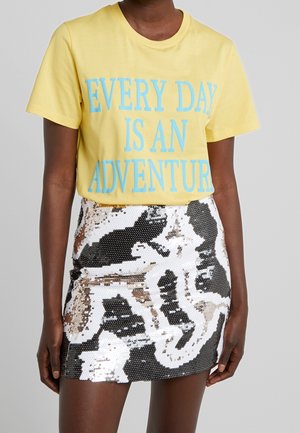 Persoon draagt een geel T-shirt met blauwe tekst "EVERY DAY IS AN ADVENTURE" en een zwarte, witte en zilveren paillettens minirok.