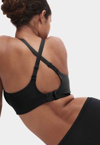 Frau trägt einen schwarzen Sport-BH mit verstellbaren, gekreuzten Trägern und Hakenverschluss, von hinten vor einem einfarbigen Hintergrund gezeigt.