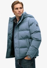 Chaqueta acolchada azul claro con capucha, con textura acolchada y cierres de presión. Se lleva sobre una camisa oscura y se combina con vaqueros.