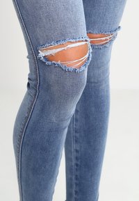 Jeans de denim azul desgastado con rodillas rasgadas, ajuste slim y bordes desflecados, que presentan un lavado claro con una textura sutil.