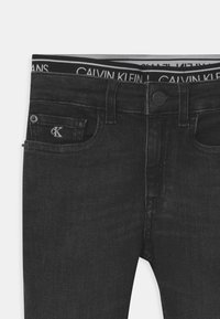 Jeans de mezclilla negra con un diseño ajustado, que cuentan con una cintura con el logo de Calvin Klein y un pequeño bolsillo con un acento de botón plateado.