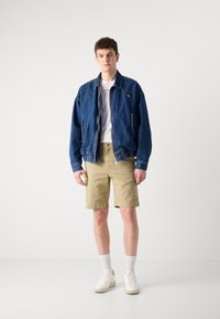 Denim jacket in dark blue with zip closure, two side pockets, and ribbed cuffs. Paired with beige cargo shorts and white sneakers.