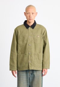 Khaki-grøn jakke med en sort fløjlskrage, frontknaplukninger og to store forlommer. Stoffet har en glat tekstur.
