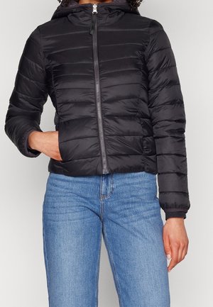 Persoon draagt een zwarte doorgestikte jas met rits en capuchon, en een blauwe spijkerbroek, hand in de jaszak.