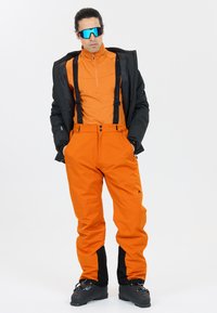 Pantalons de ski orange vif avec bretelles noires réglables, boutons frontaux et accents noirs aux chevilles, associés à une veste isolée noire et des lunettes de soleil.