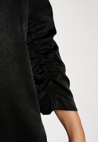 Manche de robe noire avec détail de tissu froncé au-dessus d'un avant-bras nu sur un fond clair uni.