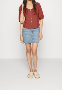 Rostfarbenes kurzärmliges Hemd, kombiniert mit einem hellen Jeansrock mit fransigem Saum und beigen Slippers. Eine cremefarbene Handtasche ist vorhanden.