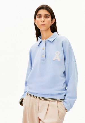 Frau mit langen dunklen Haaren trägt einen hellblauen, geknöpften Sweatshirt mit weißem Logo und beige plissierte Hosen mit Händen in den Taschen.