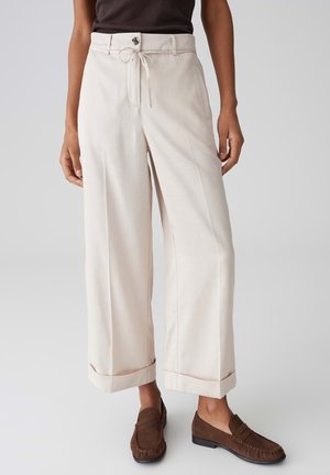 Donna che indossa pantaloni beige a gamba larga e cropped con orli risvoltati, una coulisse annodata in vita e mocassini marroni, in piedi su uno sfondo grigio.