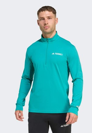 Turkusowa longsleeve z zamkiem błyskawicznym na ćwierć i beżowym logo. Posiada teksturowane boczne panele oraz dopasowane rękawy do użytku sportowego.