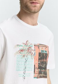 Hvid bomulds t-shirt med et strålende palme-træ grafik og tekst, samt et farvefotoelement i koral- og blå nuancer på fronten.
