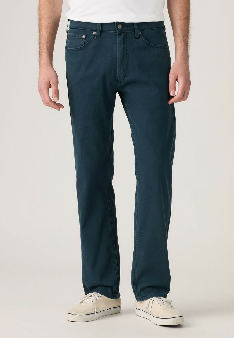 Mann trägt dunkelblaue Jeans mit geradem Bein und weiße Turnschuhe, steht vor neutralem Hintergrund.