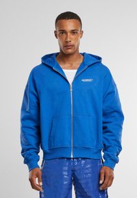 Blaue Zip-Hoodie aus weichem Stoff, mit einer Kängurutasche und einer Kapuze mit Kordelzug. "RETENRITY"-Logo auf der Brust.