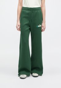 Άτομο με σκούρο πράσινο φαρδύ παντελόνι φόρμας με κορδόνι στη μέση και λευκά αθλητικά παπούτσια με μαύρα λογότυπα Nike.