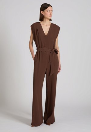 Mujer de pie con las manos en los bolsillos, vestida con un mono marrón sin mangas, con escote en V y cinturón anudado, mirando hacia un lado frente a un fondo liso.