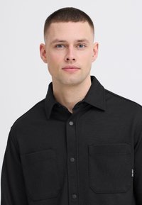 Joven con el cabello corto que lleva una camisa negra abotonada con dos bolsillos en el pecho, mirando hacia adelante contra un fondo claro y sencillo.