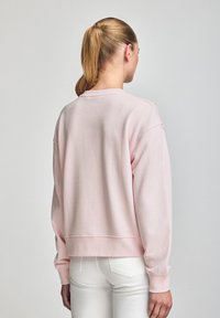 Light pink sweatshirt with a relaxed fit, ribbed cuffs, and hem, featuring a crew neckline and soft textured fabric. Worn with white jeans.