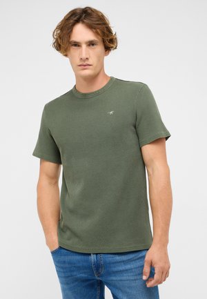 Green waffle-textured T-shirt with short sleeves and a round neckline, featuring a small logo detail on the chest. Paired with blue jeans.