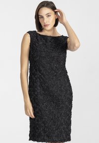 Kobieta z krótkimi, brązowymi włosami, ubrana w bezrękawnikową, strukturalną czarną sukienkę do kolan, pozująca z jedną ręką blisko twarzy.