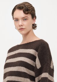 Young woman with short wavy hair wearing silver hoop earrings and a brown and beige striped knit sweater, posing against a white background.