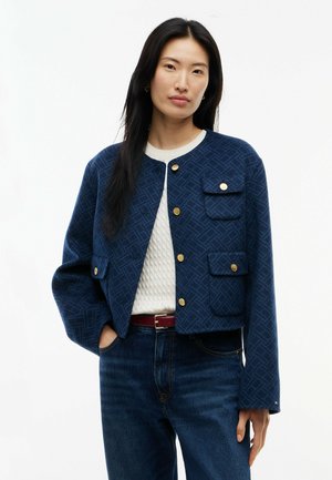 Mujer con chaqueta azul marino con estampado y botones dorados sobre un suéter blanco texturizado y vaqueros azules, de pie con una mano en el bolsillo.