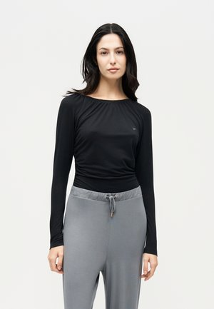 Une femme pose avec un haut noir à manches longues et un pantalon gris à cordon de serrage, sur un fond clair et uni.
