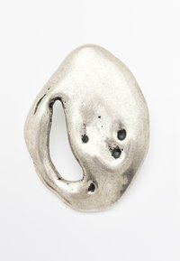 Zilverkleurige metalen broche met een organische, abstracte vorm, voorzien van een gat en drie kleine indeukingen. Glad oppervlak met een gepolijste afwerking.