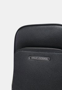 Zaino in pelle nera con finitura texturizzata, hardware argentato e tasca anteriore con zip. Presenta l'etichetta del marchio "Armani Exchange."