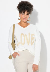 Maglione bianco a V con la scritta "LOVE" beige, abbinato a pantaloni beige chiaro. La modella indossa grandi orecchini a cerchio dorati e porta una borsa beige.