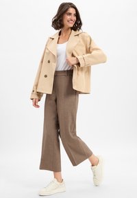 Beige dubbelzijdig jasje, brede bruine broek en off-white sneakers. Glad textuur, nauwsluitende pasvorm en contrasterende knopen.