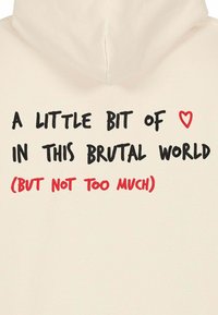 Béžová mikina s kapucňou, na ktorej je čierny a červený text: "Trochu ♥ v tomto brutálnom svete (ale nie príliš moc)" na chrbte. Mäkka tkanina.