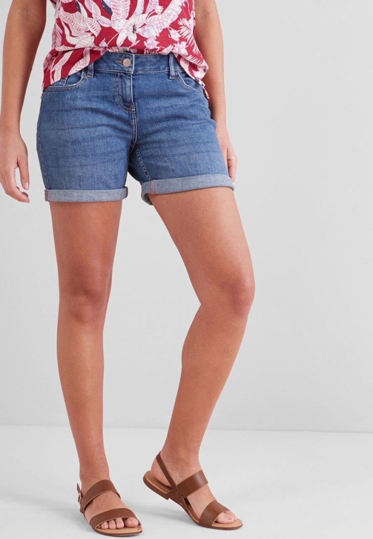 Femme portant un short en jean bleu à ourlet roulé, un haut rouge et blanc à motif, et des sandales plates marron, debout devant un fond uni.