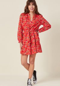 Punainen kukallinen mekko, jossa on pitkät hihat, vyötärölle sidottava nauha ja kerroksellinen hame. Mekossa on tulppaanikuvio ja se on yhdistetty mustiin lenkkareihin.