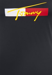 T-shirt nera con un audace logo cursivo giallo "Tommy", arricchita da un rettangolo bianco e un quadrato rosso come sfondo. Tessuto morbido e liscio.