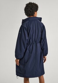 Osoba s krátkými kudrnatými vlasy, oblečená v dlouhém tmavě modrém kabátu s nařaseným pasem a elastickými manžetami, pohledem zezadu.