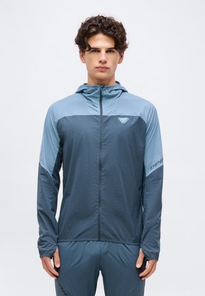 Jeune homme portant une veste à capuche zippée bicolore bleue et un pantalon de sport assorti, debout devant un fond blanc uni.