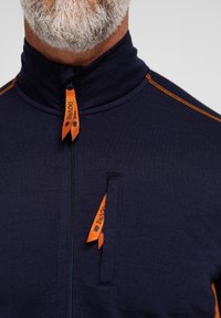 Marineblå beklædning med en høj krave, udstyret med orange syninger og lynlåstræk. Inkluderer en lille lynlåslomme på fronten.