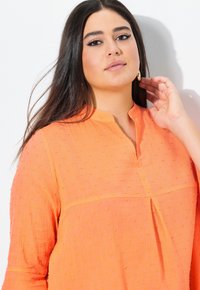 Blusa arancione brillante con un motivo testurizzato, caratterizzata da un colletto corto e una sottile piega frontale. Maniche lunghe e tessuto leggero.